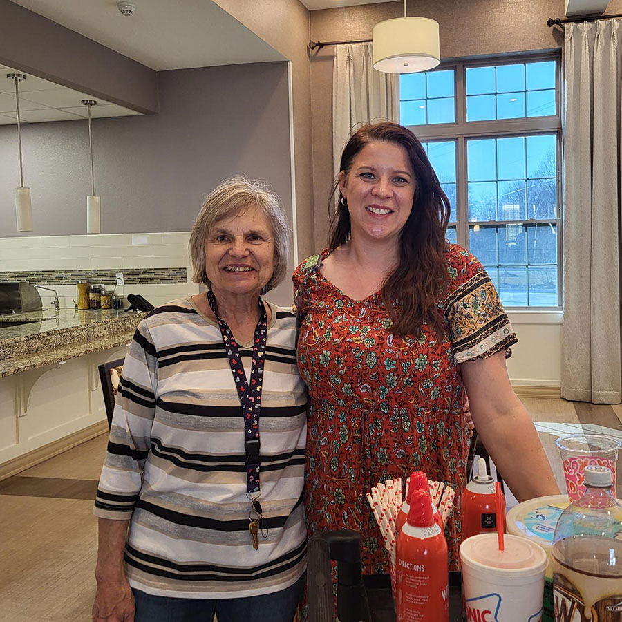 Two women smiling, standing by a table filled with food. Volunteers prepare rootbeer floats for memory care residents.