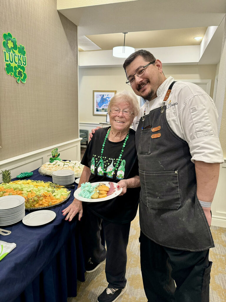 A smiling senior living chef and a resident happily selecting appetizers at a St. Patrick's Day event.