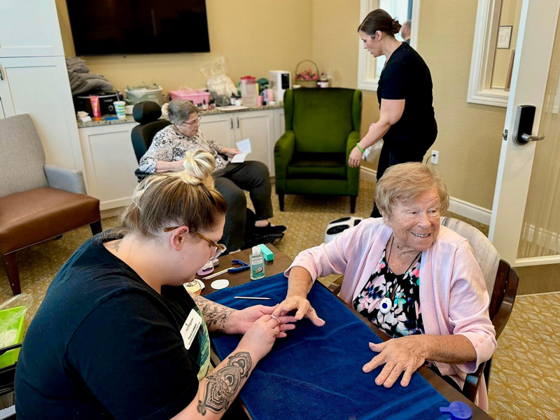 A senior resident enjoys a spa day while getting her nails done by a staff member. Another resident and staff member are in the background in a cozy setting.