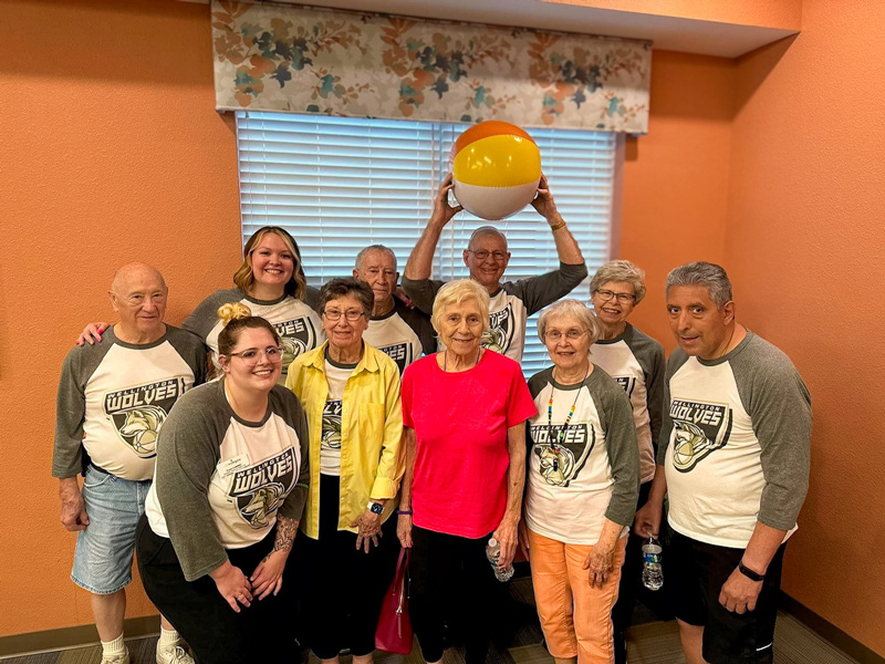 A group of senior residents and staff members pose for a fun photo after a chair volleyball game. One holds a beach ball above their head, and many wear matching team shirts.