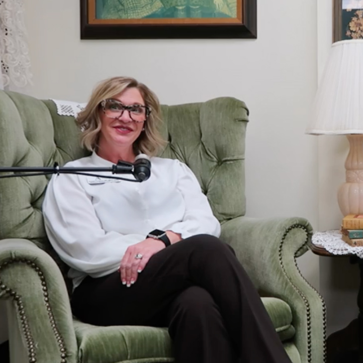 Podcast guest seated in a vintage green armchair, smiling as she speaks into a microphone for the 'Thoughtful Connections' podcast. A cozy table with books and a lamp is beside her.