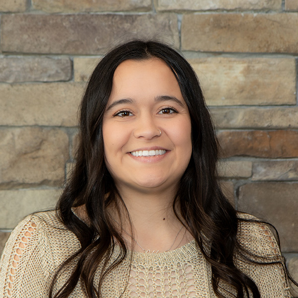 Sydney McCall, Resident Service Director at The Wellington Senior Living, smiling warmly in front of a stone wall, wearing a beige knit sweater with wavy dark hair.