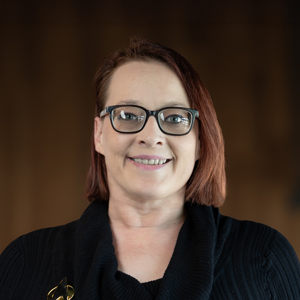 Jessica 'Jess' Jennings, Resident Services Director at The Wellington Senior Living, wearing glasses and a black sweater, smiling in a professional portrait with a warm-toned background.