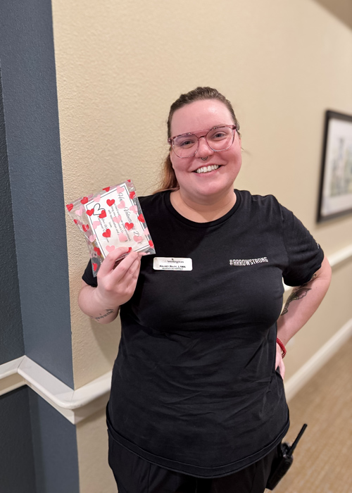 Senior living employee wearing a black shirt with #ArrowStrong printed on it, smiling while holding a heart-patterned gift bag during Employee Appreciation Day.