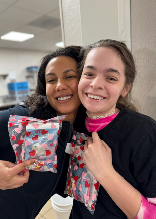 Two senior living employees pose together, smiling and holding heart-decorated gift bags as they celebrate Employee Appreciation Day with appreciation and camaraderie.