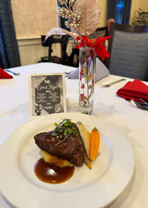 Elegant plated steak dinner with demi-glace, mashed potatoes, heirloom carrots, and microgreens, set on a white tablecloth with themed décor and menu card.