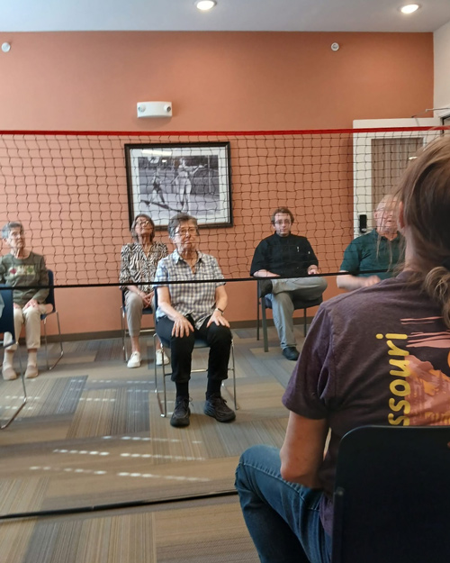 Group of senior residents participating in a chair volleyball game, seated in chairs with a net in the middle, focusing on the ball during the indoor match.