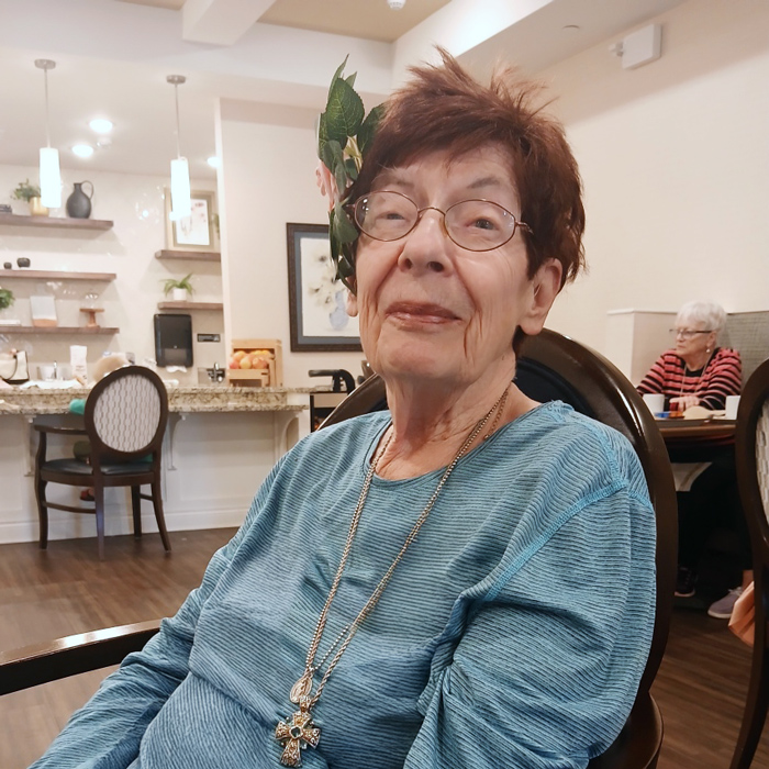 Senior resident wearing a blue top and floral hair accessory, smiling warmly while sitting in a senior living dining area.
