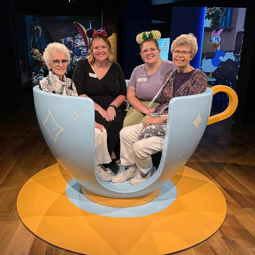 Senior residents and team members smile while seated in a large teacup at a cartoon exhibit.