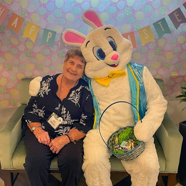 A senior resident sits on a couch with the Easter Bunny, smiling and surrounded by Easter decorations.