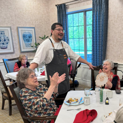 A Culinary Director shares laughter with senior residents at a dining table, creating a warm and joyful atmosphere during mealtime in the community.