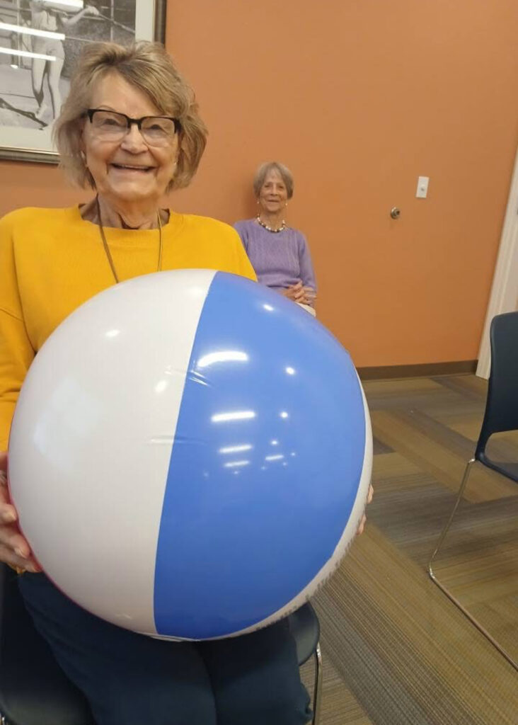Senior women in the fitness center smile, one holding a beachball.
