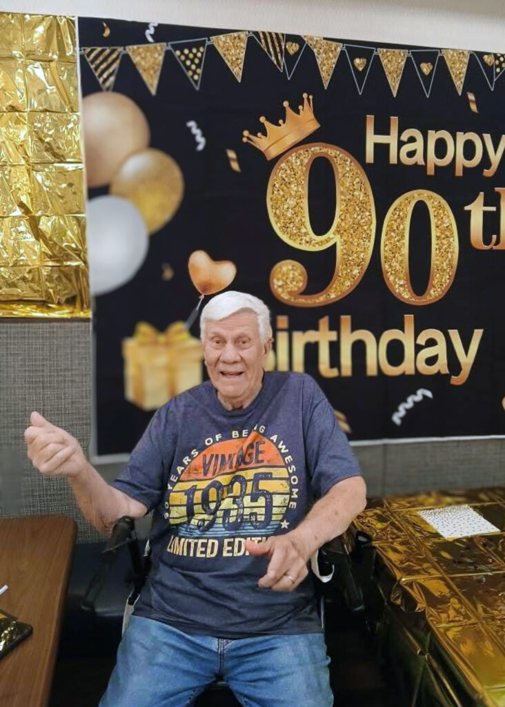 A senior man smiles in front of a birthday backdrop celebrating his 90th birthday.