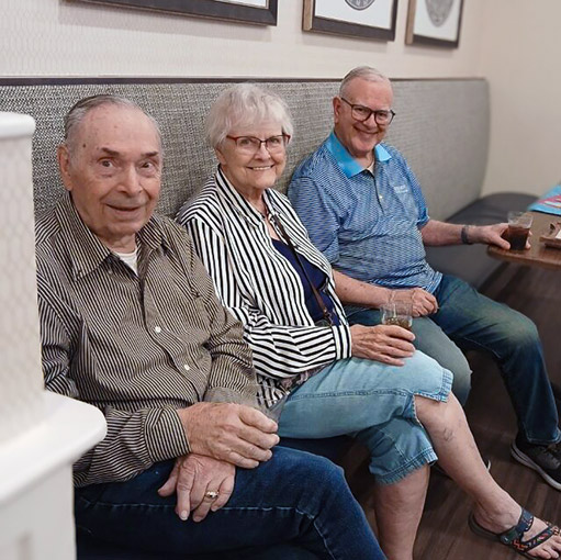 Three senior residents smile while sharing a comfortably cushioned booth at The Wellington Senior Living.