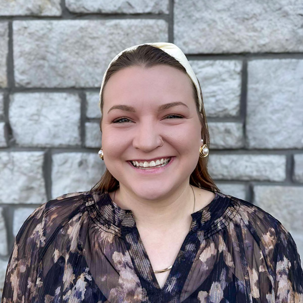 Rachel Schuckman, Resident Services Director at The Wellington Senior Living, smiling in a professional headshot, wearing a patterned blouse and headband, posed against a light stone wall.