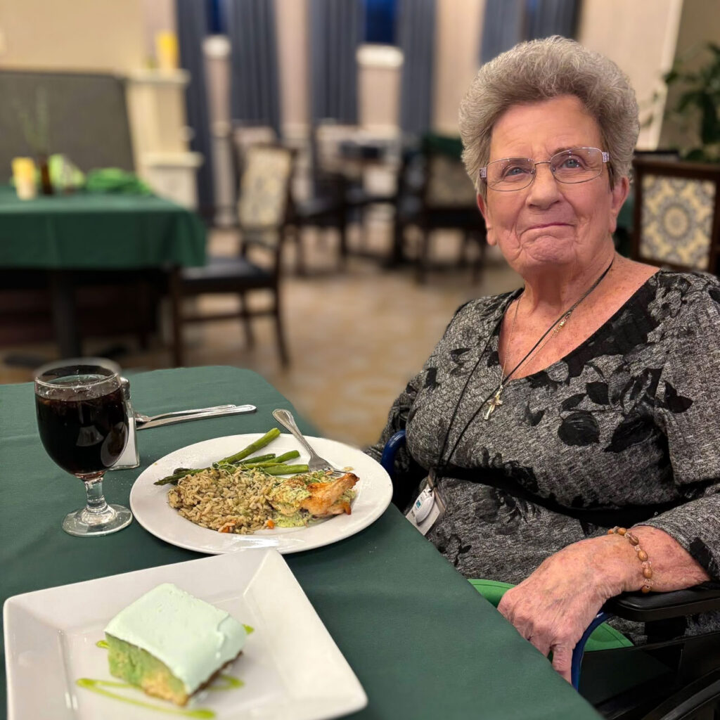 A senior woman smiles at an elegant dining table, a chef prepared meal and dessert before her.