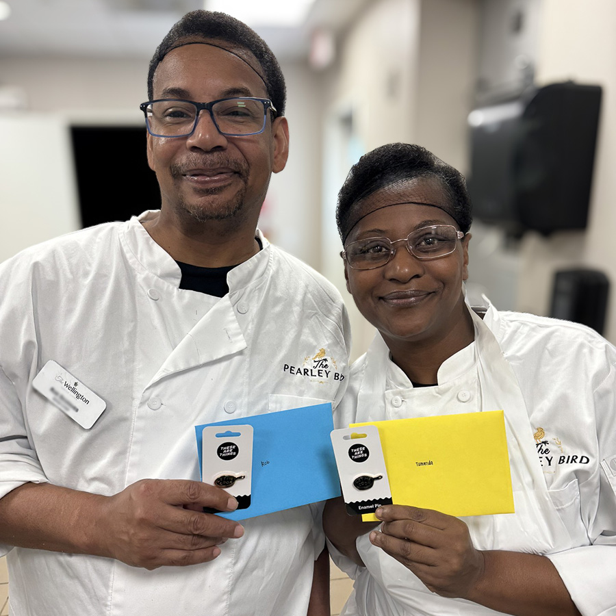 Two culinary team members at The Wellington Senior Living smile while displaying cards and skillet-themed enamel pins given in gratitude.