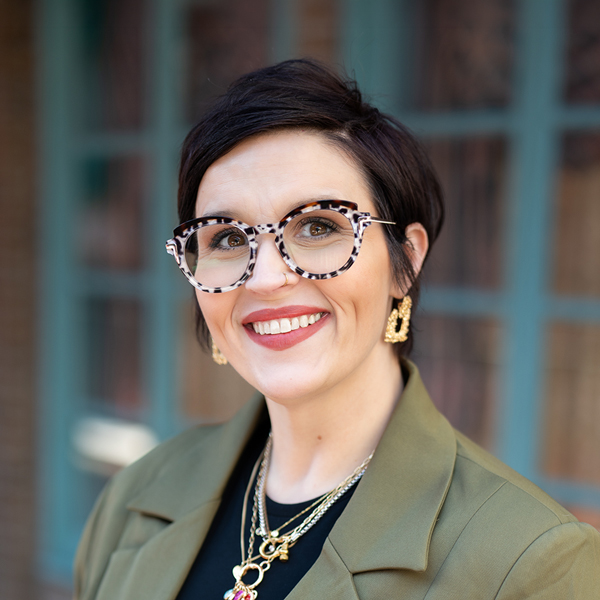Joanna Wesley, Executive Director at The Wellington Senior Living, smiling in a professional outdoor headshot with short dark hair, patterned glasses, gold jewelry, and softly blurred teal window details in the background.
