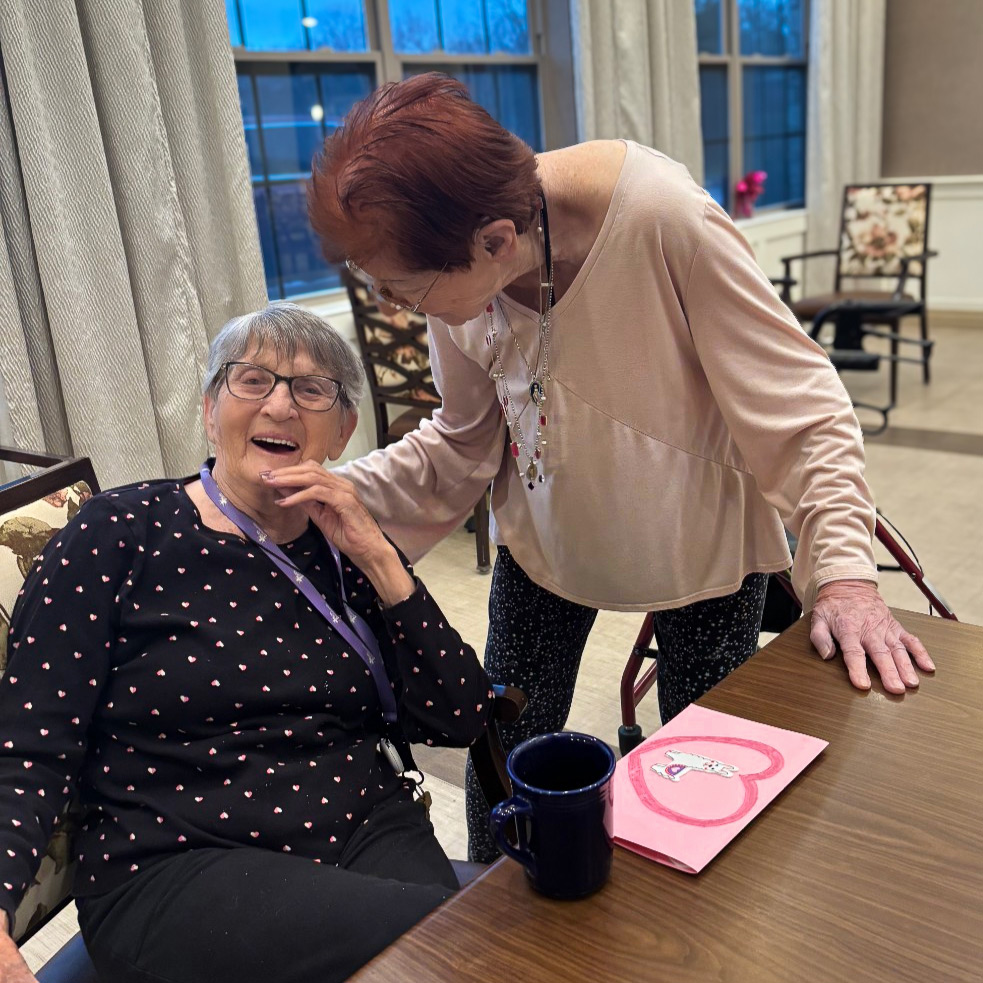 Two senior women smile together during a relaxed moment in a community common area.