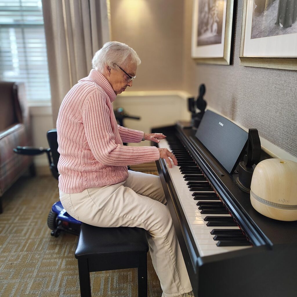 A senior woman wearing a soft pink sweater plays the piano at The Wellington Senior Living.