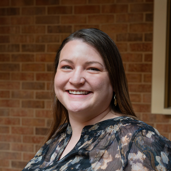 Rachel Schuckman, Resident Services Director at The Wellington Senior Living, smiling in a professional outdoor headshot with long straight dark hair, gold earrings, a patterned blouse, and a softly blurred brick background.