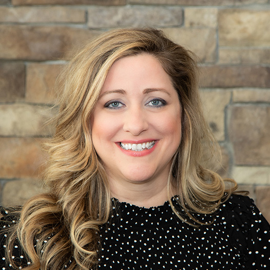 Nicole Waugh, Executive Director at The Wellington Senior Living, smiling in a professional headshot with long wavy blonde hair, a black dotted blouse, and a stacked stone wall background.