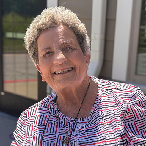 A senior woman smiles welcomingly at The Wellington Senior Living on a bright, sunny day.