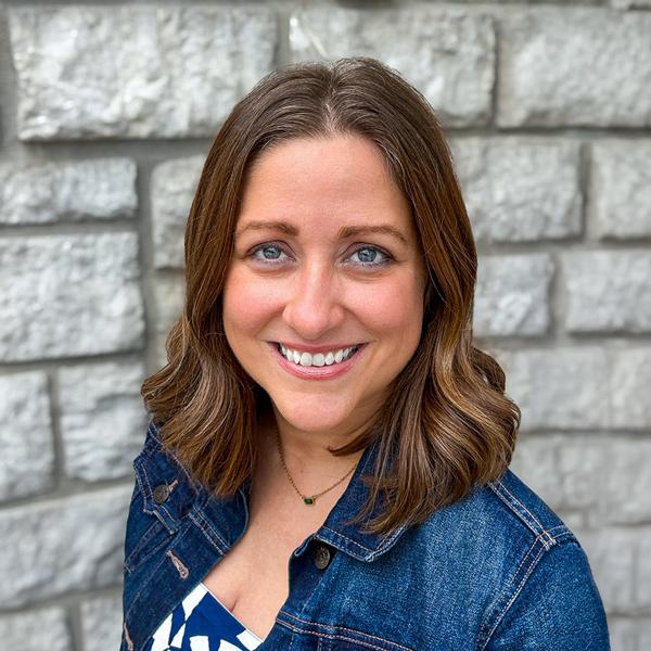 Nicole Waugh, Executive Director at The Wellington Senior Living, smiling in a professional headshot with shoulder-length wavy brown hair, a denim jacket, and a light gray stone wall background.